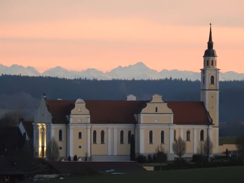 Sießener Kirche: Im Vordergrund ist die Längsseite einer Kirche zu sehen. Die Fassade ist gelb/weiß gehalten. Am rechten Ende befindet sich der etwa doppelt so hohe Kirchturm mit rundem Dach, auf dem sich dann nochmals eine Spitze erhebt. Im Hintergrund ist ein Wald zu sehen, hinter dem dann Berge folgen. Das Bild wurde in Abenddämmerung aufgenommen. 