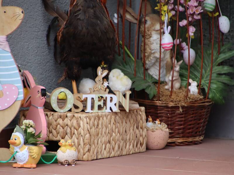 Viele Osterdekoelemente. Links ein großer Hase aus Holz, Enten und Eier aus Keramik, in der Mitte eine Kiste aus Bast auf dem ein Schriftzug OSTERN steht. Rechts davon weitere Pflanzen und ein Korb aus Bast. 