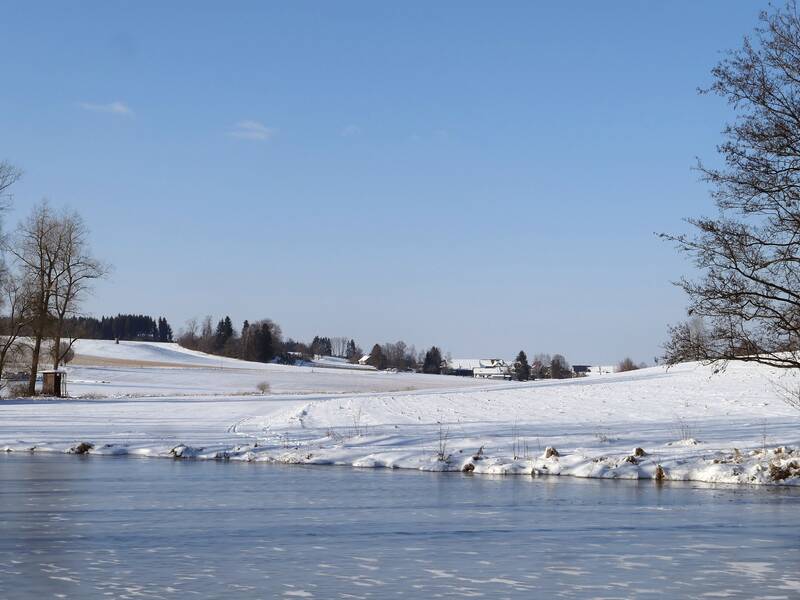 zugefrorener See im hinteren Bildbereich ist das andere Ufer mi Bäumen und einem Jägerstand zu sehen. In der Ferne ist die Siedlung Huggenlaubach zu sehen.