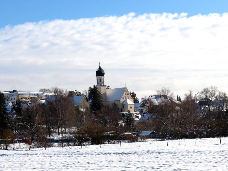 Bild einer Winterlandschaft im Vordergrund eine schneebedeckte Wiese, dahinter eine Reihe Büsche und Bäume und dahinter einige Gebäude in verschiedenen Größen, unter anderem eine Kriche mit einem Zwiebelturm.