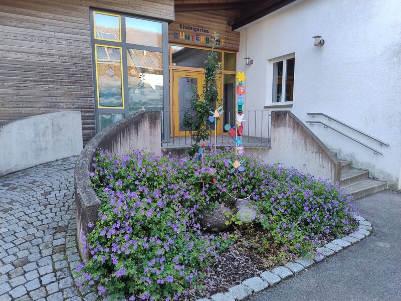 Eingangsbereich davor ein Blumenbeet bepflanzt mit Storchenschnabel. Darin sind einige bunte Gartenstecker.