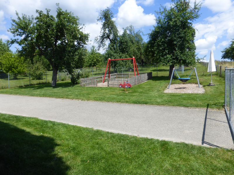 Kindergarten Außenbereich. Grüne Wiese durch einen geteerten Weg getrennt. Im Hintergrund Spielgeräte.