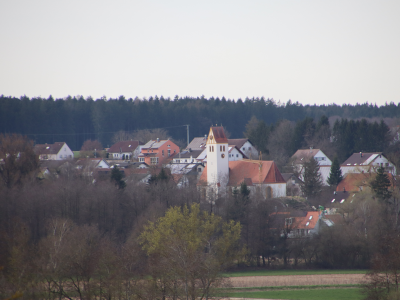 Blick auf Bußmannshausen von Großschafhausen aus. In der Bildmitte ist der Kirchturm zu sehen. Vor dem Kirchturm ist eine Baumreihe zu sehen. Um den Kirchturm herum sind weitere Gebäude zu sehen. 