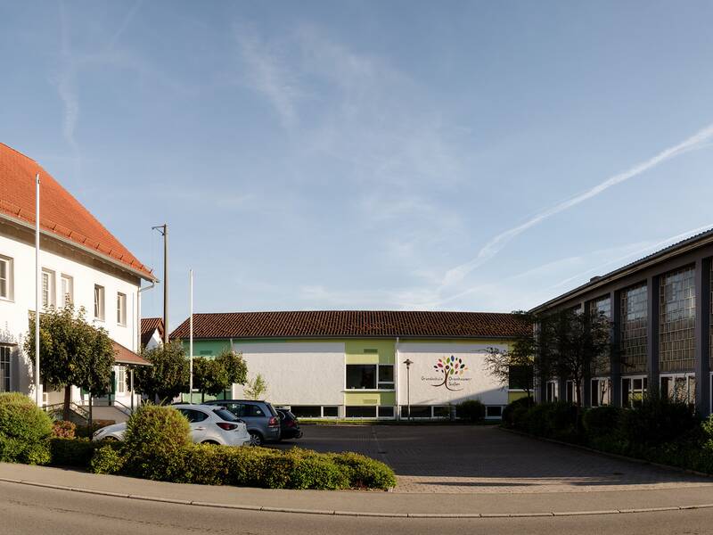 Panoramabild auf dem man links ein dreistöckiges Gebäude sieht. in der mitte ein flaches lang gezogenes Haus und Rechts eine Sporthalle.