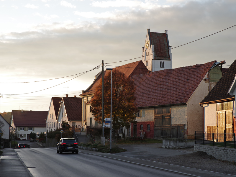 Bild entlang einer Straße. Die Straße quert das Bild von unten rechts nach oben links. Links und rechts der Straße sind viele alte Bauernhäuser. Auf der Straße fährt ein schwarzes Auto. auf der rechten Straßenseite sieht man einen Kirchturm hinter anderen Häusern hervorragen.