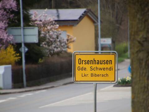 Bild des Ortsschild von Orsenhausen von Großschafhausen kommend. Im Hintergrund ist die Straße zu sehen. Links im Hintergrund sind Verteilerschränke, eine Hecke und ein blühender Baum zu sehen. Rechts wird das Bild von einem Baumstamm begrenzt.