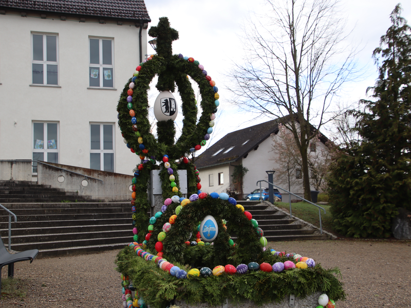 Mit Reißig und gefärbten Eiern geschmückter Brunnen vor einer Treppe. Am Ende der Treppe ist ein Haus zu sehen. An der Hausfassade sind 6 Fenster zu sehen. 