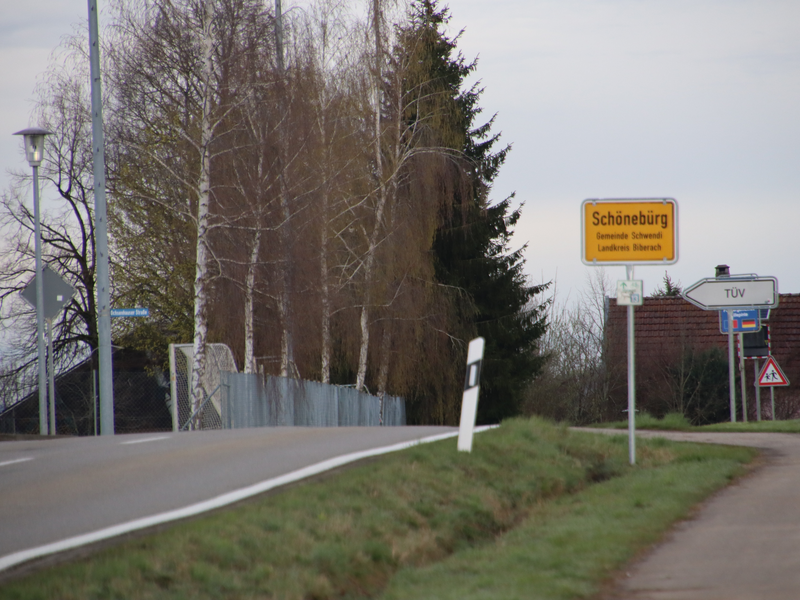 Ortseingang von Schönebürg von Hürbel kommend. links läuft die Straße, rechts ein Radweg. Die beiden asphaltierten Wege sind durch einen Grasstreifen getrennt auf dem das gelbe Ortsschild an einem metallenen Masten steht. Das Schild lautet: "Schönebürg, Gemeinde Schwendi, Landkreis Biberach". Im Hintergrund sind links Birken und Fußballtore zu sehen. rechts im Hintergrund sind weitere Verkehrsschilder zu sehen. 