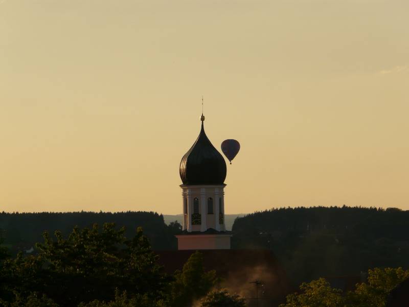 Turm der St. Stephnus Kirche in Schwendi ragt über Hausdächer,  im Hintergrund fliegt ein Heßluftballon