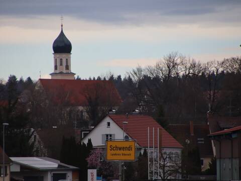 Blick auf den Ortseingang von Schwendi von Schönebürg her kommend. In der Bild Mitte ist das Gelbe Ortsschild zu sehen auf dem steht: "Schwendi Landkreis Biberach". im Hintergrund sind viele Bäume, mehrere Gebäude und ein Kirchturm zu sehen. 