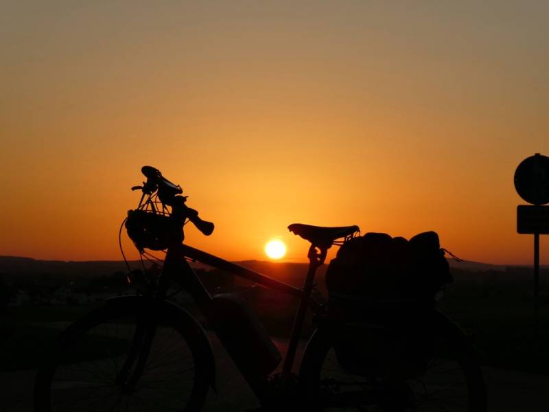 Im Vordergrund ist ein E-Fahrrad zu sehen, am Lenker hängt der Helm, am Gepäckträger sind Taschen montiert. Im Hintergrund ist die untergehende Sonne zu sehen.