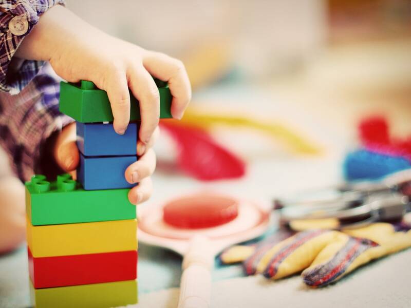 auf der linken Seite ist eine Kinderhand zu sehen, die Plastikbausteine aufeinandersetzt. rechts davon sind weitere Spiezeuge verschwommen zu erahnen. 