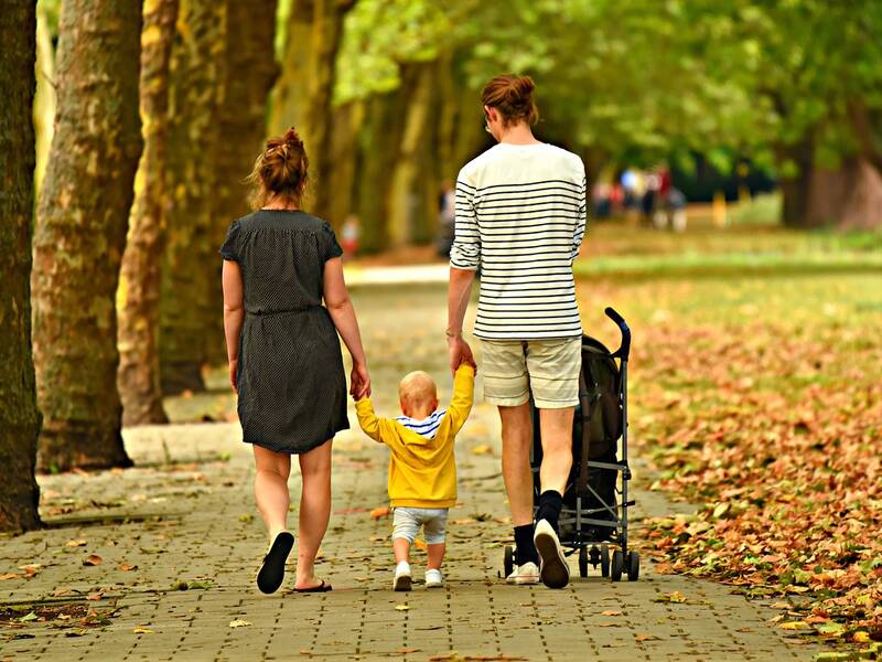 links ist eine Reihe Baumstämme zu sehen. Diese entspriesen Löchern in einem Gepflasterten Weg,. Auf dem Weg gehen eine Frau in einem Kleid (links) und ein Mann mit kurzer Hose und gestreiftem Tshirt (rechts) der einen Kinderwagen schiebt. Zwischen den beiden geht ein kleines Kind, welches von den Erwachsenen an den Händen gehalten wird. Rechts von der Szene ist eine Fläche die mit Laub bedeckt ist zu sehen. 