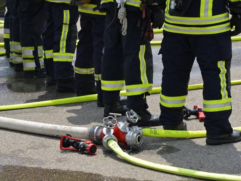 im Vordergrund sind Feuerwehrschläuche und ein Verteilter zu sehen. Dahinter stehen merhere Feuerwehrleute von denen lediglich die Beine zu sehen sind