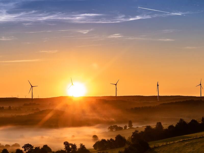 Merhere Windräder vor einem Sonnenuntergang , vor den Windkraftanlagen sind mehrere Täler zu sehen, die mit Bäumen und Nebel gefüllt sind. 