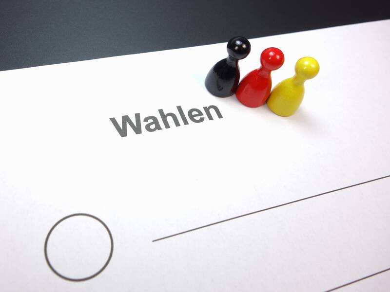 Auf einem Weißen Blatt Papier vor schwarzem Hintergrund steht das Wort Wahlen, darunter ein Kreis zum Ankreuzen. Rechts neben dem Kreis sind Linien abgebildet. Neben dem Schriftzug Wahlen stehen drei Spielfiguren in den Farben Schwarz, Rot und Gelb.