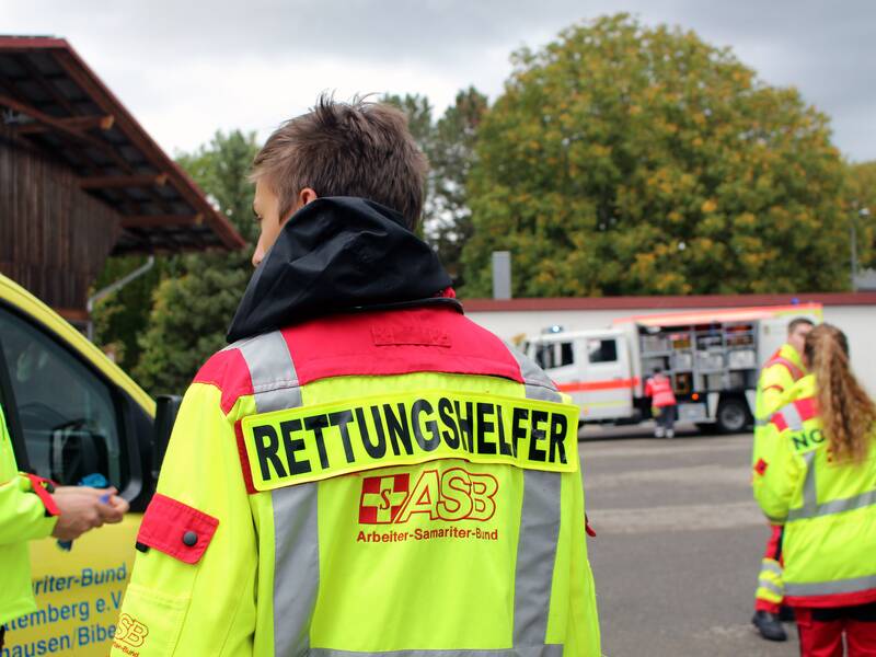 in der Mitte des Bildes ist ein Junge mit verwuschelten Haaren zu sehen, er trägt eine neongelbe Jacke mit roten Aplikationen an den Armen und auf den Schultern. Auf dem Rücken ist der Schriftzug Rettungshelfer und ASB - Arbeiter-Samariter-Bund zu lesen. Im Hintergrund sind weitere gleich gekleidete Personen und Rettungsfahrzeuge zu sehen.