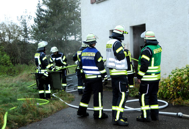 Vor einem älteren Gebäude stehen mehrere Feuerwehrmänner in Schwarz-gelber Einsatzkleidung. Die drei Personen im Vordergrund tragen bunte Warnwesten. Am Boden liegen mehrere Schlauchleitungen