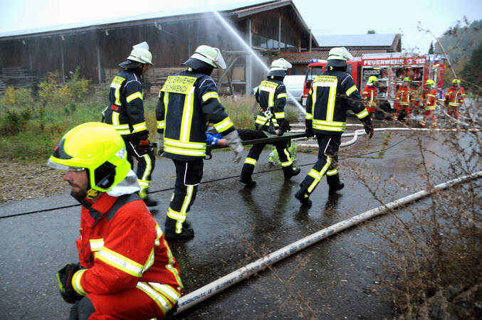 Feuerwehrmann kniet an einem Verteiler. Im Hintergrund tragen vier weitere Feuerwehrleute eine Trage in Richtung eines Feuerwehrfahrzeug.