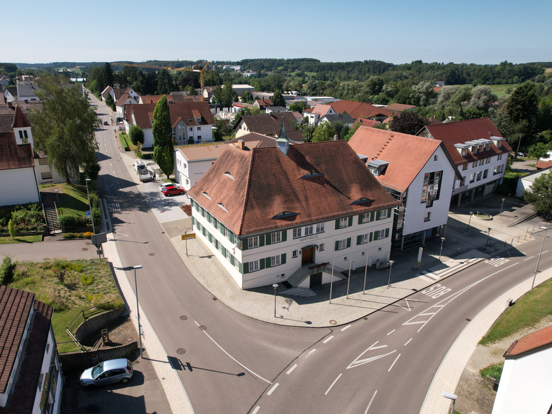 Luftbild: Blick auf das Rathaus der Gemeinde Schwendi von Norden. Im Vordergrund ist eine Straßenzweigung zu sehen (links in Richtung Gutenzell, rechts in Richtung Schönebürg). zwischen den beiden Straßen ist das Rathaus zu sehen. links der historische Altbau und rechts der angebaute Neubau. Im Hintergrund sowie links und rechts sind weitere Gebäude zu sehen.