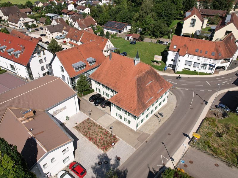 Luftbild: Blick auf das Rathaus der Gemeinde Schwendi von Süden. Im Vordergrund links ist die "Kleine Bühne" zu sehen. Rechts davon das Rathaus. zwischen den Gebäuden sind Parkplätze zu sehen. Im Hintergrund sowie links und rechts sind weitere Gebäude zu sehen.