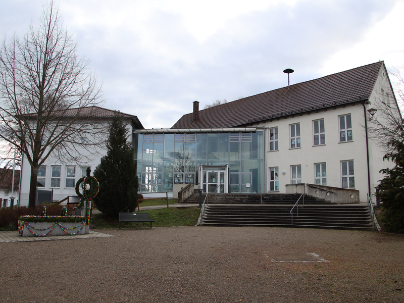 Blick auf die Grundschule in Schönebürg vom Vorplatz aus. im Vordergrund ist ein Platz zu sehen, der mit Kies bedeckt ist. Dahinter das Gebäude, welches links und rechts verputze Fassaden mit vielen Fenstern hat, welche in der Mitte mit einem Glasbau verbunden sind. Zwischen Platz und Gebäude ist eien breite Treppe zu sehen. Auf dem Kiesplatz steht noch ein kleiner Brunnen.