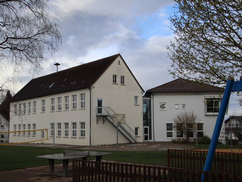 Blick auf die Rückseite der Grundschule in Schönebürg. Im Vordergrund sind mehrere Spielgeräte auf einer Grasfläche zu sehen. Dahinter erhebt sich das weiß verputze Gebäude. 