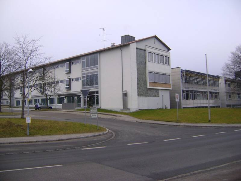 Bild der Grund- und Werkrealschule Schwendi. Auf das Gebäude wird aus Nord-Westlicher Richtung geblickt. Im Vordergrund ist die Hauptstraße zu sehen. Dahinter die Auffahrt auf den Parkplatz und dahinter das Schulgebäude. Der rechts zu sehenden neuer Teil ist mit Metall verkleidet. Die Stirnseite des Altbaus, die links direkt an den Neubau anschließt besteht aus einer Glasbausteinwand und weiteren Fenstern. Weiter links ist die Längsseite des Altbaus zu sehen, welcher ebenfalls grau/weiß ist und viele Fenster beinhaltet. 