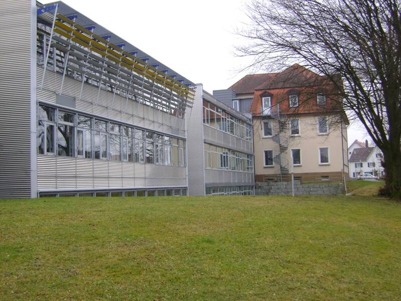 Bild der Grudn- und Werkrealschule. Im Hintergrund ist das alte Schulgebäude zu sehen. Das Rote Dach hat mehrereFenster. links davon ist die Längsseite eines neueren mit Metall verkleideten Gebäudeteil zu sehen. Davor eine Wiese. 