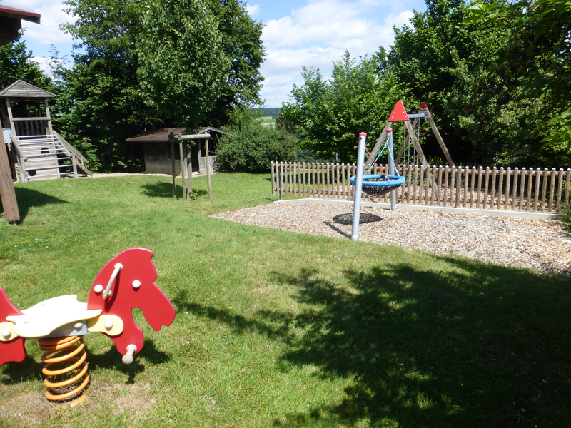 Bild von Kinderspielgeräten auf einer WIese. Sowie eine Schaukel auf einem Bereich mit Rindenmulch. Links im HIntergrund ist ein Klettergerüst zu sehen.