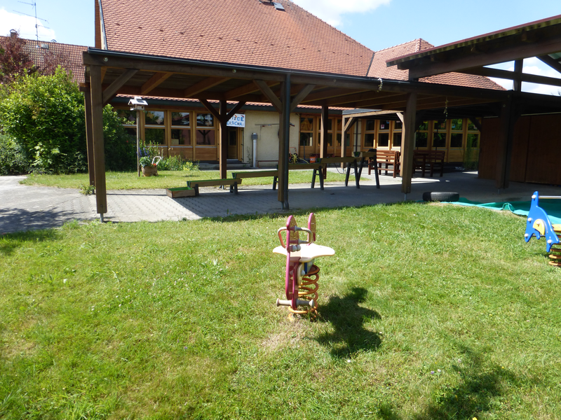 Im Hintergrund ein Haus vom dem man das Rote Ziegeldach sieht. Davor ein überdachter Platz mit Kinderspielgeräten. Davor eine WIese mit Schaukelpferden.