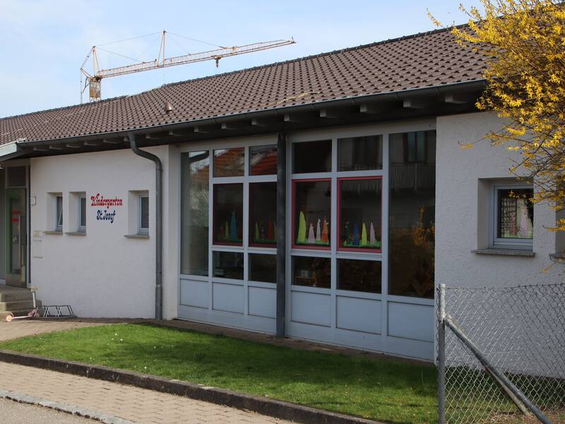 Bild von der Fassade des Kindergarten in Orsenhausen. Links ist der Eingangsbereich zu sehen. Rechts eine Fensterfront. Dazwischen ist an der Wand der Schriftzug "Kindergarten St. Josef" zu lesen. Rechts von der Fensterfront steht ein Busch mit gelben Blüten.