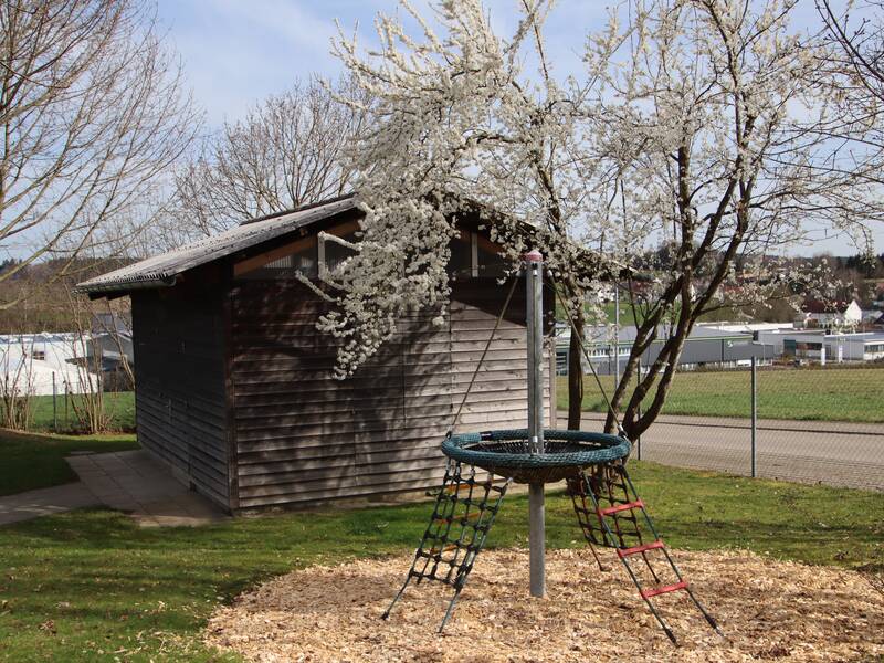 Bild des Gartens. Im Hintergrund eine Gerätehütte, davor ein Baum mit Blüten und davor ein Klettergerüst welches auf Holzschnitzeln steht.