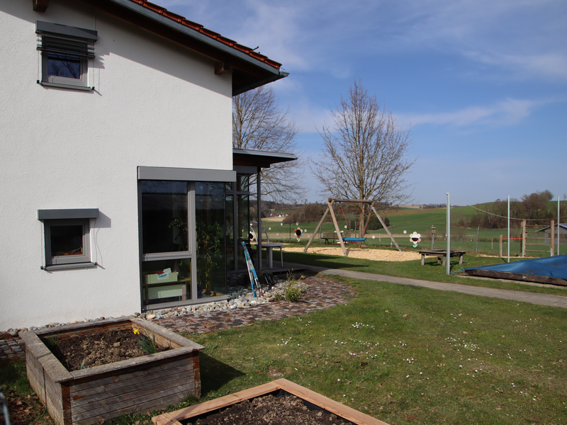 Bild des Gartens. Links ist das Gebäude des Kindergartens zu sehen. An der Fassade sind zwei Fenster und eine Fensterfront zu sehen. Vor der Fassade sind zwei Hochbeete zu sehen, rechts davon eine große Grasfläche auf der sich diverse Spielgeräte und ein Sandkasten befinden.