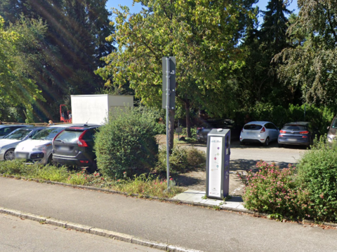 Bild von einem Parkplatz, darauf sind hinter einer Straße und einer Buschreihe mehrere Autos zu sehen. zwischen den Büschen ist eine E-Ladesäule zu sehen.