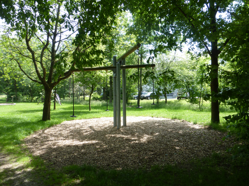 Im Schatten von merheren Bäumen auf einer Wiese ist eine Fläche mit Rindenmulch zu sehen. Darauf steht eine Hochwippe.