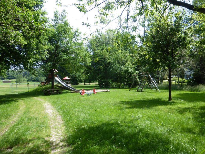 Auf einer Grünen Wiese stehen mehrere Bäume. Dazwischen sind mehrere Spielgeräte zu sehen. Eine Wippe, eine Schaukel, eine Rutsche und ein Kletterturm. Außerdem mehrere Sitzgelegenheiten.