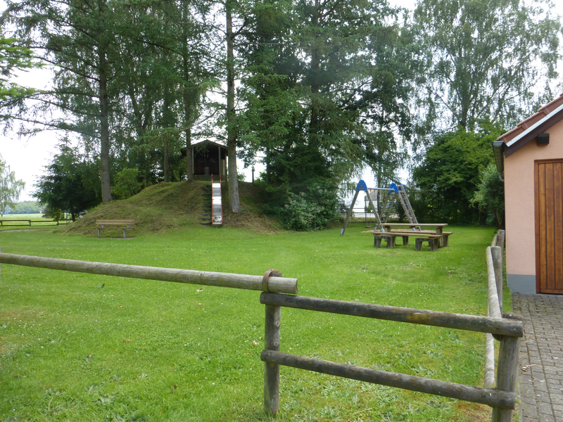 Auf einer Wiese ist rechts eine Sitzbank mit Tisch zu sehen. Dahinter eine Schaukel. Im linken Bereich ist ein Hügel zu sehen, von dem eine Rutsche nach unten führt. 