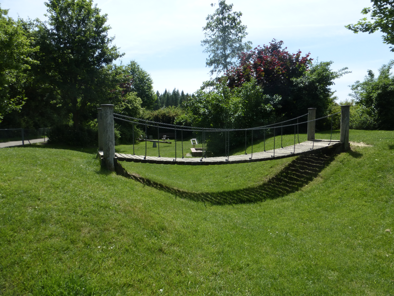 Auf einer Wiese zwischen zwei kleinen Hügeln ist eine Hängebrücke aus Holz zu sehen,