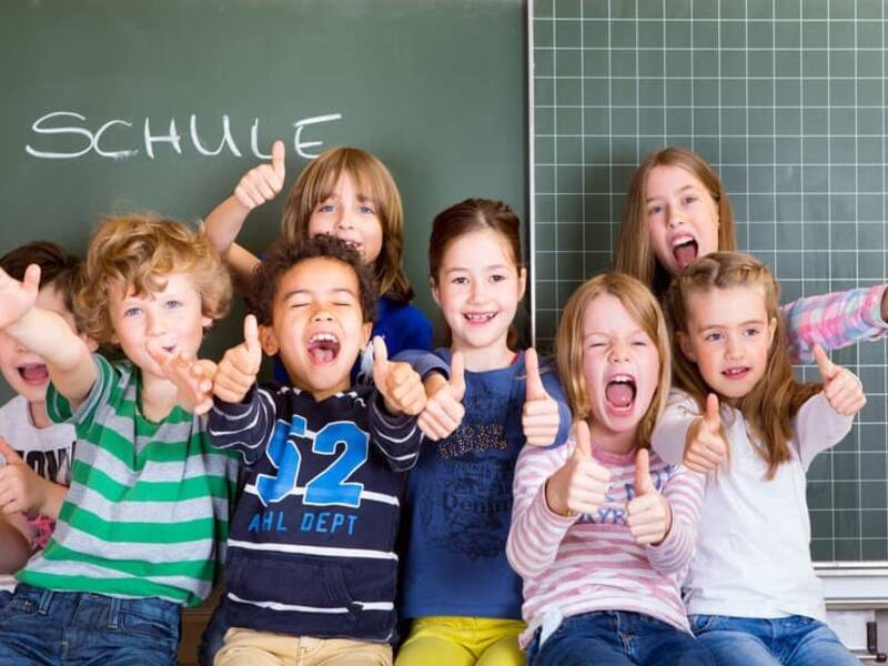 8 Kinder vor einer Tafel auf der mit weißer Kreide "Schule" geschrieben steht. Die Kinder sitzen vermutlich auf einem Tisch und strecken alle den Daumen nach oben.