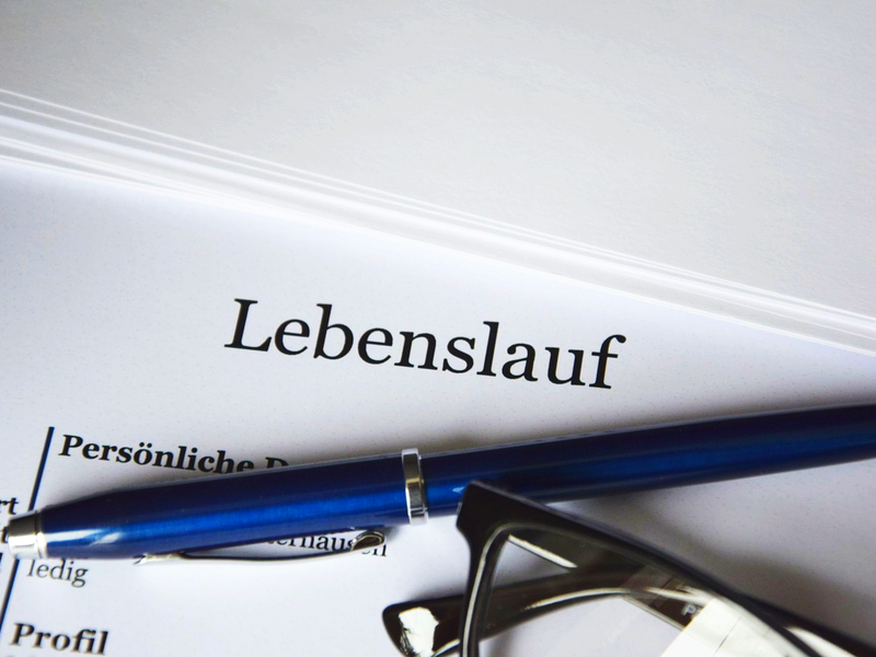 Auf einem weißen Blatt, dass auf einem weißen Tisch liegt steht in schwarzen Buchstaben "Lebenslauf". Auf dem Blatt liegt eine schwarze Brille und ein blauer Kugelschreiber