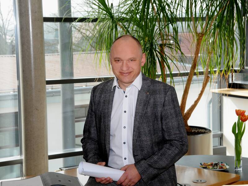 Auf einem Bürotisch sitzt ein Mann mit weißem Hemd und grauem Sakko gekleidet. Er schaut direkt in die Kamera und hält Papiere in der Hand. 