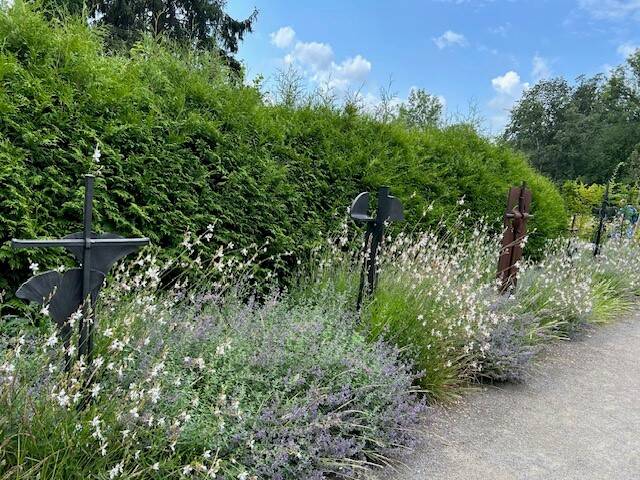 Eine Reihe von dekorativen schwarzen und rostfarbenen Metallkreuzen steht in einem Gartenpfad zwischen blühenden Pflanzen und hohen Sträuchern.